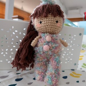 May include: A crocheted doll with brown hair, wearing a pink and blue fuzzy outfit. The doll has a pink nose and rosy cheeks.