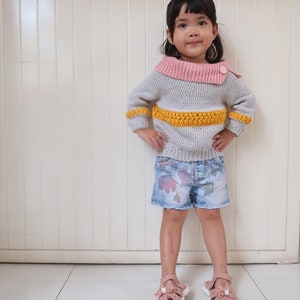 Puede incluir: Una niña pequeña con un suéter gris, rosa y amarillo a rayas con un cuello rosa y un solo botón. Lleva pantalones cortos de mezclilla azul con estampado floral y sandalias rosas.