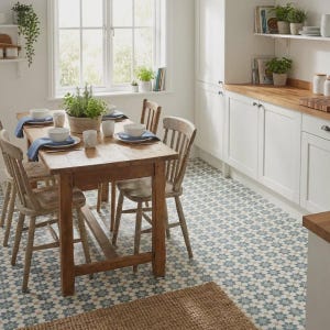May include: A rustic kitchen scene featuring a wooden table set for a meal, surrounded by wooden chairs. White cabinets and a window with natural light create a bright atmosphere. The floor has a patterned tile design, and a woven rug is in the foreground.