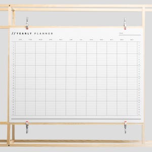 Könnte beinhalten: Ein weißer Jahresplaner mit einem schwarzen Raster und den Worten "// YEARLY PLANNER" oben. Der Planer hängt an einem Holzrahmen.