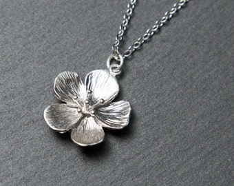Puede incluir: Un collar de plata con un delicado colgante de flor. La flor está detallada con pétalos texturizados y un pequeño centro.
