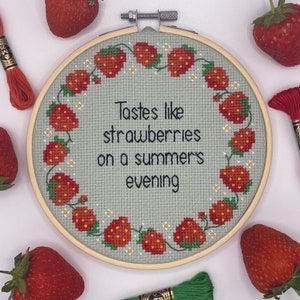 May include: A round embroidery hoop with a light blue background and a white border. The hoop features a red and green embroidered design of strawberries and vines. The text "Tastes like strawberries on a summer's evening" is embroidered in black thread.