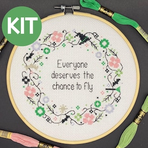 May include: Cross-stitch kit with a floral wreath design and the text "Everyone deserves the chance to fly." The embroidery hoop is wooden, and the kit includes green and pink embroidery floss. A green circle with the word "KIT" is in the upper left corner.