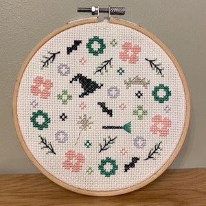 May include: A circular cross-stitch embroidery, featuring a witch's hat, bats, flowers, and other Halloween motifs. The design is stitched on white fabric and set in a wooden hoop.