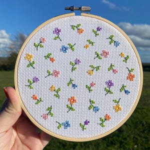 Könnte beinhalten: Ein runder Stickrahmen aus Holz mit weißem Aida-Stoff, der ein Kreuzstichmuster aus kleinen, verstreuten Blumen in Rosa, Gelb, Orange, Lila und Blau zeigt, wobei jede Blume zwei kleine grüne Blätter an einem dünnen grünen Stiel hat.