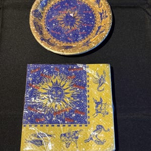 May include: A gold-rimmed plate and a stack of napkins featuring a celestial design. The plate has a blue center with a sun and zodiac symbols. The napkins are yellow and blue, with a sun graphic and zodiac illustrations.