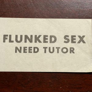 May include: A rectangular card with the words "FLUNKED SEX NEED TUTOR" printed in a bold, sans-serif font. The card is off-white and rests on a dark brown wooden surface.