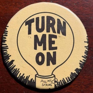 May include: A round pinback button with a light yellow background and black ink illustrations. The design features a lightbulb shape containing the text TURN ME ON in bold letters, with a small base labeled PULL MY STRING in black text.