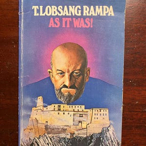 May include: A book cover with a purple background and the title "T. Lobsang Rampa As It Was!" in white text. The cover features a portrait of a man with a beard and a mountain scene in the background.