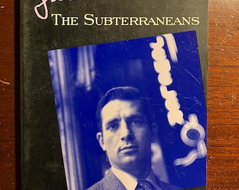 May include: A black and white book cover with the title "The Subterraneans" by Jack Kerouac. A black and white photo of a man is on the cover. The text "A book of raw power and awesome beauty." is written below the photo, with the attribution "San Francisco Examiner".