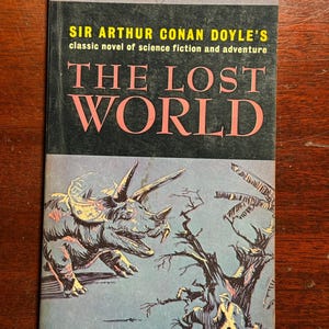 May include: A vintage paperback book cover featuring a black and yellow illustration of a dinosaur, a tree, and two people. The title of the book is "The Lost World" by Sir Arthur Conan Doyle.