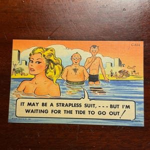 May include: Vintage postcard featuring a cartoon illustration of three people in the ocean. A woman with blonde hair and two men are in the water. The text reads, "IT MAY BE A STRAPLESS SUIT, - - - BUT I'M WAITING FOR THE TIDE TO GO OUT/"
