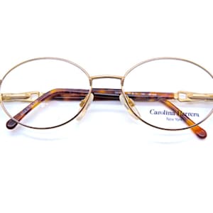 Op de afbeelding: Ronde gouden bril met schildpadkleurige pootjes en de tekst "Carolina Herrera New York" op de rechter poot.