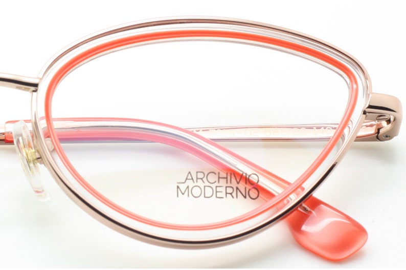 May include: Rose gold cat-eye glasses with a pink and clear frame. The brand name "Archivio Moderno" is printed on the frame.