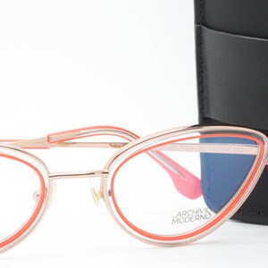 May include: Rose gold cat eye glasses with a bright orange frame. The glasses are sitting on a white surface next to a black leather glasses case with the text "Archivio Moderno" on it.