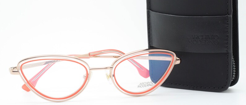 May include: Rose gold cat eye glasses with a bright orange frame. The glasses are sitting on a white surface next to a black leather glasses case with the text "Archivio Moderno" on it.