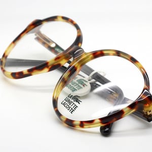 May include: Tortoiseshell-patterned eyeglasses with clear lenses. The frames have a rounded shape and a dark brown and yellow color scheme. The brand name "LA LUNETTE LACOSTE" is visible on a tag attached to the glasses.