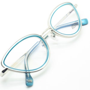 May include: Silver cat-eye glasses with blue accents. The frames have a thin silver metal band with a thicker blue band around the lenses. The brand name "ARCHIVIO MODERN" is printed on the right temple.