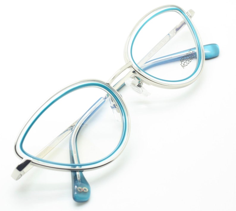 May include: Silver cat-eye glasses with blue accents. The frames have a thin silver metal band with a thicker blue band around the lenses. The brand name "ARCHIVIO MODERN" is printed on the right temple.