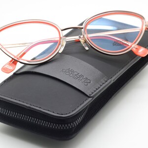 May include: A pair of pink and gold cat-eye glasses with a black leather case. The case has a zipper closure and the text "Archivio Moderno" printed on it.