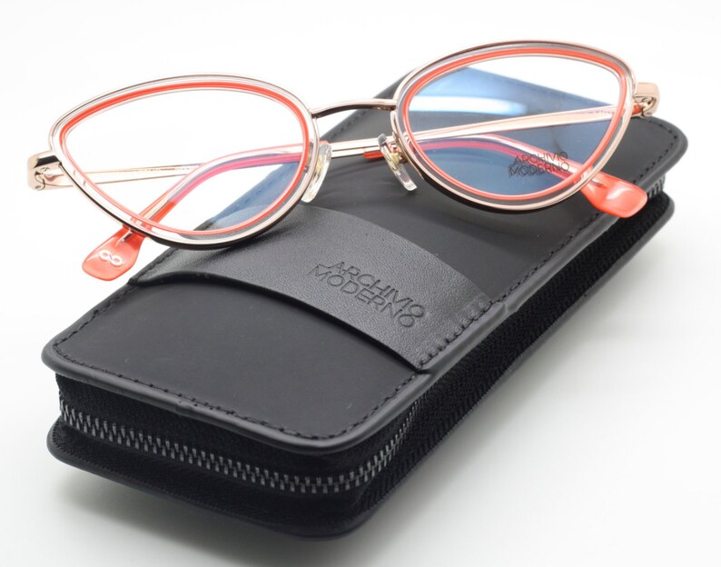 May include: A pair of pink and gold cat-eye glasses with a black leather case. The case has a zipper closure and the text "Archivio Moderno" printed on it.