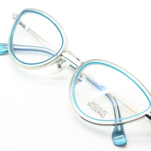 May include: Silver cat-eye glasses with a blue rim. The glasses have the text "Archivio Moderno" on the right lens.