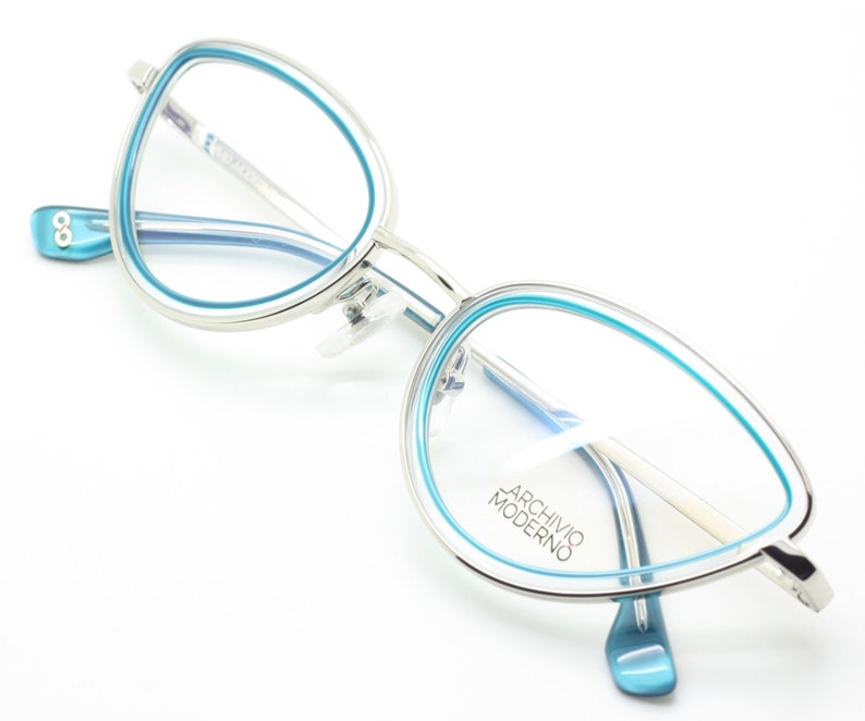 May include: Silver cat-eye glasses with a blue rim. The glasses have the text "Archivio Moderno" on the right lens.