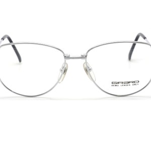 May include: Silver-toned metal eyeglasses with a cat-eye shape and clear lenses. The brand name "GIRARD" is printed on the right lens with the text "DEMO LENSES ONLY".