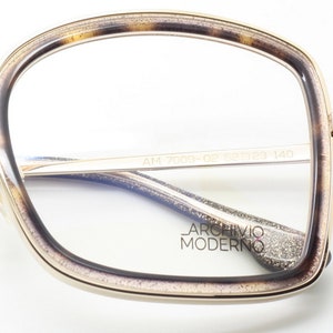 May include: Gold-rimmed eyeglasses with a tortoise shell frame and a sparkly, textured finish. The glasses have a blue tint and the brand name "Archivio Moderno" is printed on the arm. The model number "AM 7009-OP 55□123 140" is printed on the arm.
