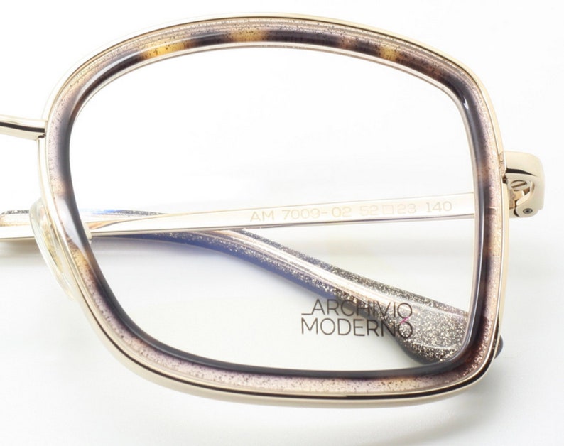 May include: Gold-rimmed eyeglasses with a tortoise shell frame and a sparkly, textured finish. The glasses have a blue tint and the brand name "Archivio Moderno" is printed on the arm. The model number "AM 7009-OP 55□123 140" is printed on the arm.