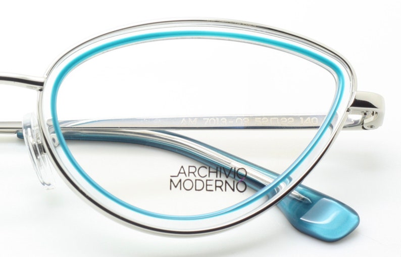 May include: Silver eyeglasses with a blue rim and a blue accent on the temple. The brand name "Archivio Moderno" is printed on the lens. The glasses have the model number "AM 7013-02 52□19 140" printed on the temple.