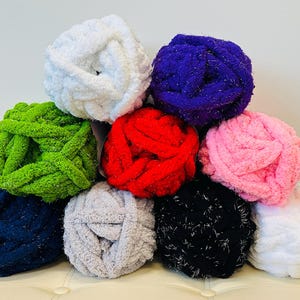 Puede incluir: Una colección de ovillos de lana coloridos en varios tonos, incluyendo blanco, verde, rojo, rosa, negro y azul. La lana parece suave y gruesa, adecuada para proyectos de tejido o ganchillo. Los ovillos están apilados en forma de pirámide.