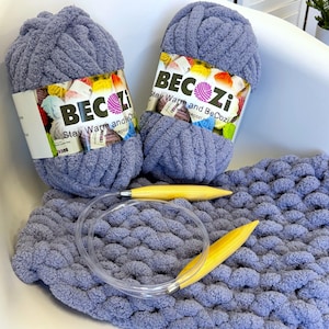 May include: Two skeins of chunky, periwinkle blue yarn with white labels reading BECOZI Stay Warm and BeCozi, resting on a partially knitted blanket of the same yarn, accompanied by a pair of large, yellow circular knitting needles.