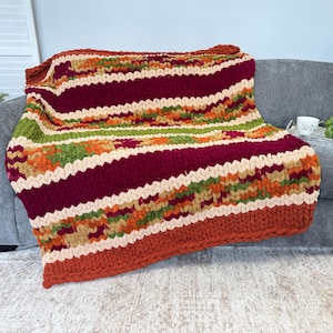 Op de afbeelding: Een grof gebreide plaid met horizontale strepen in donkerbordeaux, cr&egrave;me, olijfgroen en gebrand oranje. De deken heeft een dikke, getextureerde garenconstructie met een zichtbaar gevlochten breipatroon over het gehele oppervlak.