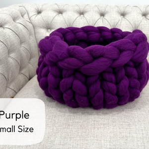 May include: A deep purple basket crafted from thick, chunky yarn in a braided knit pattern. The basket features a round, open-top design with a soft, textured appearance. A white label in the corner displays the text Purple Small Size.