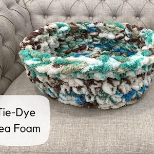 Puede incluir: Una cama redonda para mascotas te&ntilde;ida con tintes en colores espuma de mar. La cama est&aacute; hecha de un material suave y texturizado con tonos de blanco, azul, marr&oacute;n y verde. El texto "Tie-Dye Sea Foam" est&aacute; en un cuadro blanco.