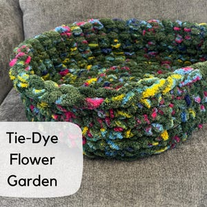Puede incluir: Una cesta te&ntilde;ida a mano con efecto tie-dye en tonos verdes, azules, amarillos y rosas. La cesta tiene forma ovalada y est&aacute; hecha de un hilo grueso y texturizado. El texto "Tie-Dye Flower Garden" se muestra en una etiqueta blanca.