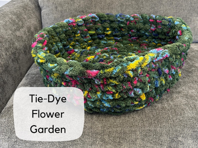 Puede incluir: Una cesta te&ntilde;ida a mano con efecto tie-dye en tonos verdes, azules, amarillos y rosas. La cesta tiene forma ovalada y est&aacute; hecha de un hilo grueso y texturizado. El texto "Tie-Dye Flower Garden" se muestra en una etiqueta blanca.