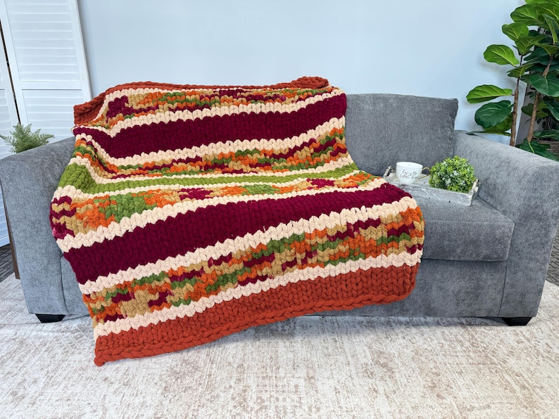 Op de afbeelding: Een grof gebreide plaid met horizontale strepen in donkerbordeaux, cr&egrave;me, olijfgroen en gebrand oranje. De deken heeft een dikke, getextureerde garenconstructie met een zichtbaar gevlochten breipatroon over het gehele oppervlak.
