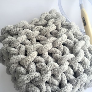 Peut inclure: Une couverture en tricot épaisse de couleur grise, réalisée avec un fil épais. La couverture est réalisée avec un motif de point simple et a une texture douce et duveteuse.