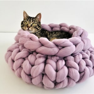 May include: A round, chunky knit pet bed made from thick, soft lavender yarn. The bed features a braided, oversized stitch pattern that creates a deep, circular basin. A brown tabby cat with green eyes is resting inside the lavender yarn bed.