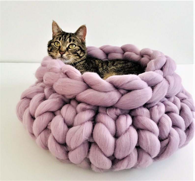 May include: A round, chunky knit pet bed made from thick, soft lavender yarn. The bed features a braided, oversized stitch pattern that creates a deep, circular basin. A brown tabby cat with green eyes is resting inside the lavender yarn bed.