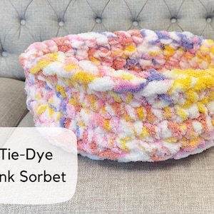 Puede incluir: Una cama redonda para mascotas tejida a mano con un patr&oacute;n de te&ntilde;ido anudado. La cama presenta una mezcla de hilo rosa, amarillo, blanco y morado. El texto "Tie-Dye Pink Sorbet" se muestra en una etiqueta blanca.