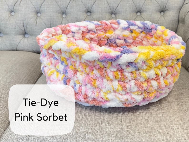 Puede incluir: Una cama redonda para mascotas tejida a mano con un patr&oacute;n de te&ntilde;ido anudado. La cama presenta una mezcla de hilo rosa, amarillo, blanco y morado. El texto "Tie-Dye Pink Sorbet" se muestra en una etiqueta blanca.