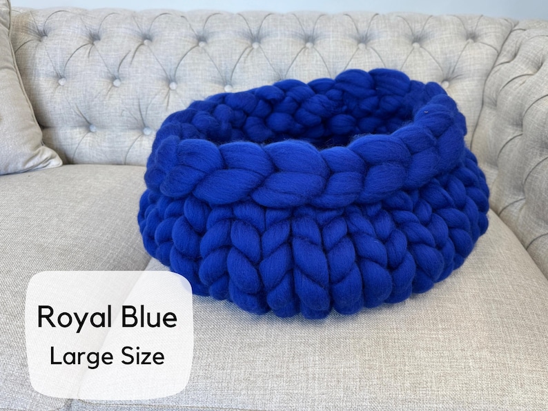 May include: A large, round, royal blue basket crafted from thick, chunky knit yarn. The basket features a prominent braided texture throughout its circular structure. A white label in the corner displays the text Royal Blue Large Size.