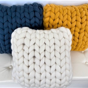 May include: Three chunky knit throw pillows in different colors. The pillows are white, mustard yellow, and dark blue. The pillows are arranged on a white couch.