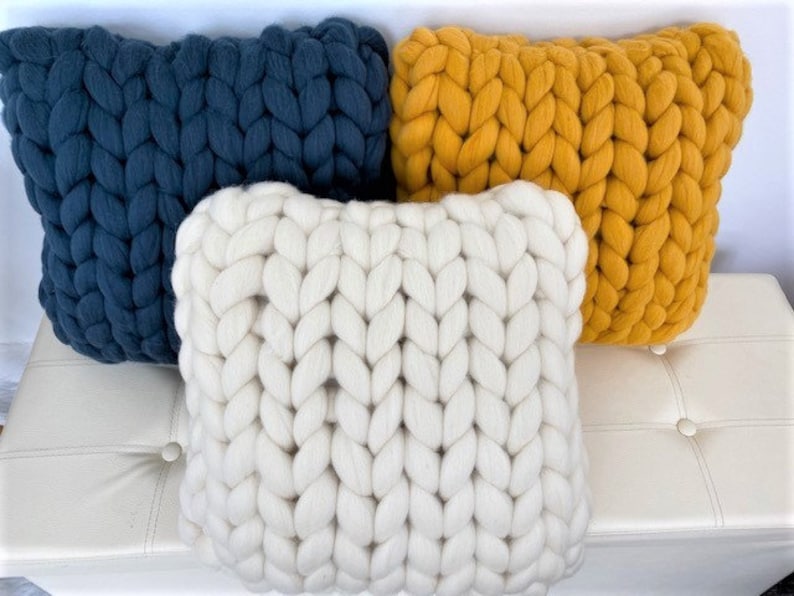 May include: Three chunky knit throw pillows in different colors. The pillows are white, mustard yellow, and dark blue. The pillows are arranged on a white couch.