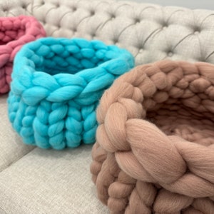May include: Three round baskets crafted from thick, chunky yarn in shades of dusty pink, bright turquoise, and soft beige. Each basket features a prominent, braided knit texture with a deep, open interior and a sturdy, rounded base structure.