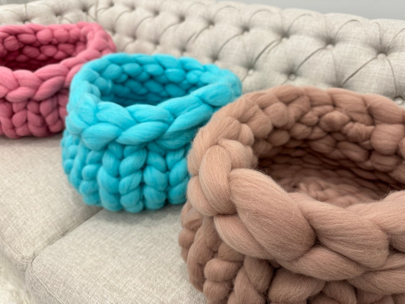 May include: Three round baskets crafted from thick, chunky yarn in shades of dusty pink, bright turquoise, and soft beige. Each basket features a prominent, braided knit texture with a deep, open interior and a sturdy, rounded base structure.