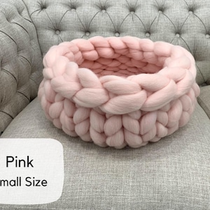 May include: A small, round, light pink basket crafted from thick, chunky knit yarn. The basket features a braided, interlocking texture throughout its circular structure. A white label in the corner displays the text Pink Small Size.